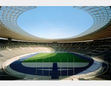 Ein großes, modernes Stadion mit einem ovalen Grundriss, umgeben von überdachten Sitzrängen. Im Zentrum befindet sich ein grünes Spielfeld, das von einer blauen Laufbahn umgeben ist.