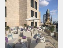 Terrasse eines Gebäudes mit modernen weißen Loungemöbeln, blauen und violetten Kissen sowie Sonnenschirmen, im Hintergrund die Turmruine der Kaiser-Wilhelm-Gedächtniskirche und weitere Stadtansichten.