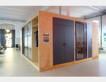 Holzverkleideter, freistehender Raum mit schwarzen Türen und Glasfenstern in einer offenen Büroumgebung mit weiß gefliesten Wänden und großzügigem Platz. Im Hintergrund sind industrielle Elemente, moderne Beleuchtung und minimalistische Möbel zu sehen.