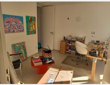 Ein heller Raum mit Atelier- oder Arbeitszimmercharakter. An der Wand hängen bunte Kunstwerke, ein Schreibtisch mit vielen Stiften und Materialien steht neben einem Regal mit Büchern und Ordnern. Kartons und ein kleiner Beistelltisch sind ebenfalls im Raum verteilt.