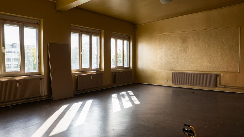 Leerer, renovierungsbedürftiger Innenraum mit gelblichen Wänden, drei großen Fenstern und Heizkörpern darunter; Sonnenlicht wirft lange Schatten auf den dunklen Boden. An der rechten Wand befindet sich eine große rechteckige Wandfläche, daneben lehnt eine Tür an der Wand.