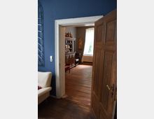 Innenansicht eines Wohnbereichs mit geöffneter Holztür, die den Durchgang zu einem Raum mit Holzdielenboden zeigt. Im Hintergrund stehen ein Holzschrank mit dekorativen Gefäßen, ein großes Fenster mit weißen Vorhängen und eine Heizung.