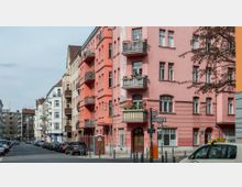 Straßenszene in einem Wohngebiet mit mehrstöckigen Altbauhäusern. Die Fassaden sind in verschiedenen Farben gestaltet, darunter ein auffälliges rosa Gebäude mit Balkonen; geparkte Autos stehen entlang der Straße, und Straßenschilder sowie ein Baum sind sichtbar.