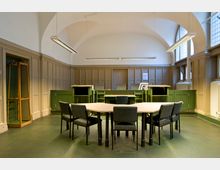 Ein historischer Versammlungsraum mit hohen gewölbten Decken, großflächigen Fenstern und Holzvertäfelung an den Wänden. In der Mitte befinden sich ein runder Tisch mit grauen Stühlen und ein grünes Richterpult im hinteren Bereich.