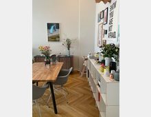 Esszimmer mit hellem Holzboden und modernen grauen Stühlen an einem rechteckigen Holztisch. Im Hintergrund stehen ein Sideboard mit Dekorationen und ein weißes Regal mit Pflanzen und Büchern; an der Wand hängen Bilder und Poster.