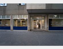 Straßenansicht eines grauen Gebäudes mit großen gläsernen Schaufensterfronten und einer zentralen Eingangstür aus Glas. Der untere Bereich der Fenster ist mit blauen Folien abgedeckt, und das Pflaster des Gehwegs davor besteht aus grauen Steinen.