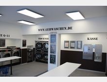 Ein moderner Waschsalon mit mehreren Waschmaschinen und Trocknern an den Wänden. An der rechten Seite befinden sich ein Kassenautomat und ein Waschmittelspender; an der Wand hängen Anleitungen und Preistafeln.