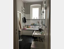 Modernes Badezimmer mit dunkelgrauem Fliesenboden, einer weißen Badewanne mit integriertem schwarzen Mosaikfliesenstreifen und einem kleinen Fenster mit weißem Rahmen. Ein weißes Waschbecken, ein Handtuchheizkörper und diverse Spielzeuge sowie Pflegeprodukte sind sichtbar.