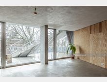 Minimalistischer Innenraum mit Betonboden und -decke, einer Wand mit Sperrholzverkleidung und einer großen Fensterfront, die den Blick auf eine Außentreppe und Bäume freigibt. Ein Topfpflanze steht als einziger Einrichtungsgegenstand in der Ecke.