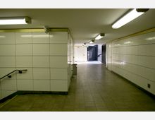 Unterführung mit weißen, rechteckigen Wandfliesen und grauem Bodenbelag. Die Decke ist mit langen, rechteckigen Deckenleuchten ausgestattet, und am Ende ist ein Ausgang mit Tageslicht und dunklen Gittertüren sichtbar.