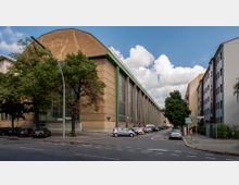 Straßenszene mit einer großen, historischen Industriehalle aus gelbem Stein mit hohen Fenstern und grünen Akzenten. Im Vordergrund eine Kreuzung mit geparkten Autos, umgeben von Wohngebäuden und Bäumen.