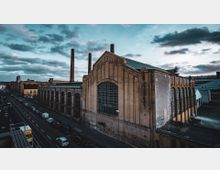 Eine historische Industriehalle aus Backstein mit großen Fenstern und hohen Schornsteinen, flankiert von einer Straße mit parkenden Autos auf der linken Seite. Im Hintergrund sind weitere Industrie- und Stadtgebäude unter einem bewölkten Himmel zu sehen.