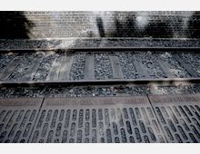 Schienen eines Bahngleises mit einem Gedenkelement aus Metall im Vordergrund, das Gravuren mit Daten, Ortsnamen und Hinweisen auf Deportationen während des Holocaust trägt. Im Hintergrund ist eine gemauerte Wand teilweise im Schatten sichtbar.