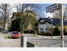Straßenszene in einer Wohngegend mit gepflastertem Gehweg, mehreren Bäumen und einem Straßenschild mit der Aufschrift "Alsenstraße". Links ein bewachsenes Mehrfamilienhaus und rechts ein freistehendes Haus mit weißer Fassade und schwarzen Fensterläden, davor parkende Autos.