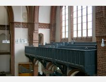 Innenansicht einer Kirche mit Backsteinwänden, Spitzbogenfenstern und einer erhöhten, hölzernen Empore in dunklem Blau. Die Szene zeigt auch eine Orgel, dekorative Holzschnitzereien und moderne, hängende Deckenleuchten.