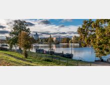 Flussufer mit Blick auf eine ruhige Wasserfläche, flankiert von Bäumen und Grünflächen im Vordergrund. Im Hintergrund sind moderne Gebäude, ein Wohnblock und ein kleines Bootsanleger zu sehen, umgeben von herbstlichem Laub und teils bewölktem Himmel.