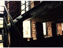 Industrie-innenraum mit roten Backsteinwänden, großen Fenstern und einer Metalltreppe. Die Konstruktion zeigt sichtbare Metallträger und Geländer, die einen industriellen Charakter hervorheben.