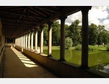 Überdachter Arkadengang mit Steinpfeilern und Holzdach, der an einem ruhigen Teich entlangführt; im Hintergrund sind Bäume und eine Wiese zu sehen.