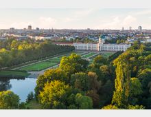 Luftaufnahme des Schlosses Charlottenburg in Berlin mit symmetrisch angelegtem Schlossgarten im Vordergrund, umgeben von vielen Bäumen; im Hintergrund eine urbane Skyline mit Wohn- und Bürogebäuden.