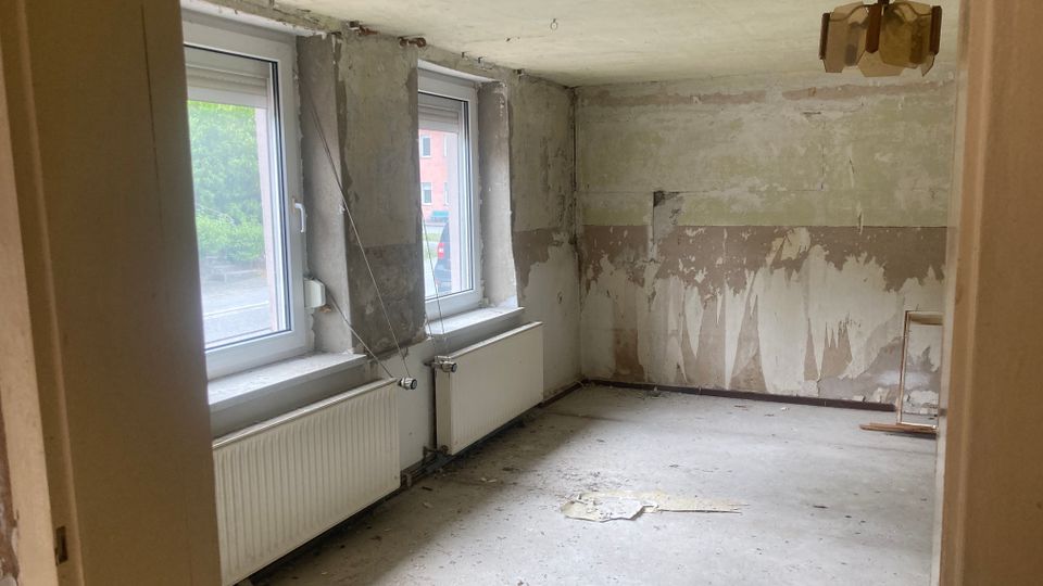 Ein leerer, renovierungsbedürftiger Raum mit zwei großen Fenstern auf der linken Seite, Heizkörpern darunter und abblätterndem Putz an den Wänden. Die Decke ist schlicht, und eine beschädigte Deckenleuchte hängt in der Mitte des Raumes.