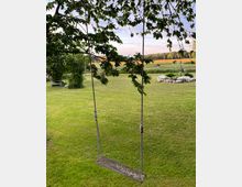 Holzschaukel an einem Baum mit Blick auf eine weitläufige Wiese, umgeben von Bäumen und Blumenbeeten; im Hintergrund Äcker und ein bewaldeter Horizont unter einem leicht bewölkten Himmel.