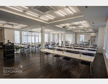 Konferenzraum mit einer modernen Einrichtung, bestehend aus langen weißen Tischen und schwarzen Stühlen in Reihen angeordnet. Der Raum hat eine helle Beleuchtung durch Deckenleuchten, große Fenster mit Jalousien und ist mit Holzfußboden ausgestattet.