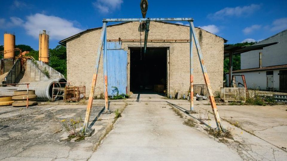 Industriegelände mit einer alten, beige-farbenen Lagerhalle, offenstehendem blauen Tor und rostigem Metallgerüst im Vordergrund. Im Hintergrund sind Betonröhren, gelbe Silos und weitere Gebäude zu sehen.