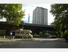 Eine städtische Szene mit einer erhöhten Bahnbrücke, unter der geparkte Autos und ein Wohnmobil stehen. Im Hintergrund steht ein hohes Wohngebäude, umgeben von Bäumen, während die gepflasterte Straße im Vordergrund liegt.