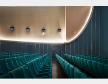 Moderner Kinosaal mit gepolsterten, türkisfarbenen Sitzreihen, die in geordneten Reihen stehen. Die Wände sind mit blauen Paneelen und goldenen Linien gestaltet, mit dezenten Wandlampen und einer geschwungenen beleuchteten Decke.