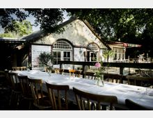 Ein rustikales Gebäude mit hell gestrichener Holzfassade, großen Fenstern mit Rundbögen und einem Dach mit Schindeloptik, umgeben von Bäumen. Im Vordergrund steht ein langer Holztisch mit weißen Tischdecken und Blumenvasen unter freiem Himmel.