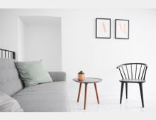 Minimalistisch eingerichtetes Wohnzimmer mit einem grauen Sofa, einem kleinen runden Beistelltisch aus Holz mit einem Kupfergefäß und einer Pflanze darauf, sowie einem schwarzen Stuhl mit geschwungener Rückenlehne. An der weißen Wand hängen zwei gerahmte Bilder mit Buchstaben.