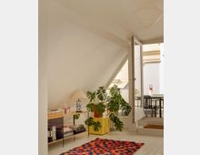 Helles Wohnzimmer mit Dachschrägen, einem farbenfrohen Teppich, einem Regal mit Büchern und Dekoartikeln, einer großen Monstera-Pflanze und einer geöffneten Tür, die zu einem Balkon mit Tisch und Stühlen führt.