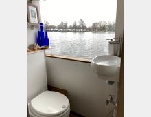Kleines Badezimmer auf einem Boot mit einer weißen Toilette, einem kleinen Waschbecken und einem Wandregal mit blauen Glasflaschen und Dekoration. Durch ein großes Fenster ist ein Blick auf einen See und ein Ufer mit Bäumen und Gebäuden sichtbar.