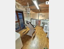 Behandlungsraum in einer Praxis mit höhenverstellbarem Stuhl in der Mitte, Holzfußboden und freiliegender Ziegeldecke mit Leuchtstofflampe. Entlang der Wände stehen Schränke und Arbeitsflächen mit Geräten sowie ein Fensterbereich mit Geländer.