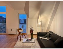 Helles Wohnzimmer in einer Dachgeschosswohnung mit schrägen Wänden, Holzboden und modernen Möbeln. Zu sehen sind ein schwarzes Sofa, ein Beistelltisch mit Büchern und Blumen, ein Holzstuhl sowie große Fenster mit Blick auf umliegende Dächer.