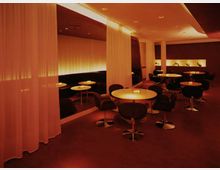 Ein modernes Lounge-Interieur mit gedämpfter, warmer Beleuchtung, runden Tischen und gepolsterten Stühlen auf einem roten Teppichboden. Der Raum ist durch halbtransparente Vorhänge und Nischen mit dekorativer Beleuchtung gestaltet.