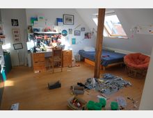 Dachzimmer mit Holzboden, eingerichtet mit einem Schreibtisch aus Holz, einem Stuhl, einem Einzelbett und einem orangefarbenen, runden Stuhl. Auf dem Boden liegen Spielzeug, hauptsächlich Lego-Steine, verstreut, und das Zimmer ist durch ein schräges Dachfenster gut beleuchtet.