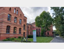 Historisches Backsteingebäude mit großen Fenstern und dekorativen Mustern, umgeben von Grünflächen und Bäumen. Im Vordergrund stehen ein Wegweiser zur Notaufnahme und ein angeschlossenes Fahrrad; rechts führt ein Gehweg entlang des Gebäudes.