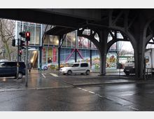 Straßenszene unter einer Eisenbahnbrücke mit Blick auf ein modernes Gebäude mit großen Glasflächen und farbigen Stahlelementen. Die Fassade ist mit bunten Graffiti verziert, und eine Rolltreppe führt ins Gebäude; im Vordergrund sind eine Ampel und vorbeifahrende Autos zu sehen.