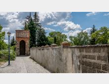 Gepflasterter Weg mit einer historischen Torbogenstruktur aus Backstein und Stein, flankiert von einer hohen Mauer und umgeben von Bäumen und Sträuchern bei sonnigem Wetter. Im Vordergrund steht eine Laterne, und der Himmel ist mit Wolken bedeckt.