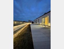 Moderne Gebäudeterrasse bei Dämmerung mit breitem Gehweg aus großen hellen Steinplatten, angrenzender Glasfassade mit beleuchtetem Innenraum und niedriger Brüstung mit Beleuchtung auf der linken Seite. Im Hintergrund sind ein Gebäude und ein bewölkter Himmel erkennbar.