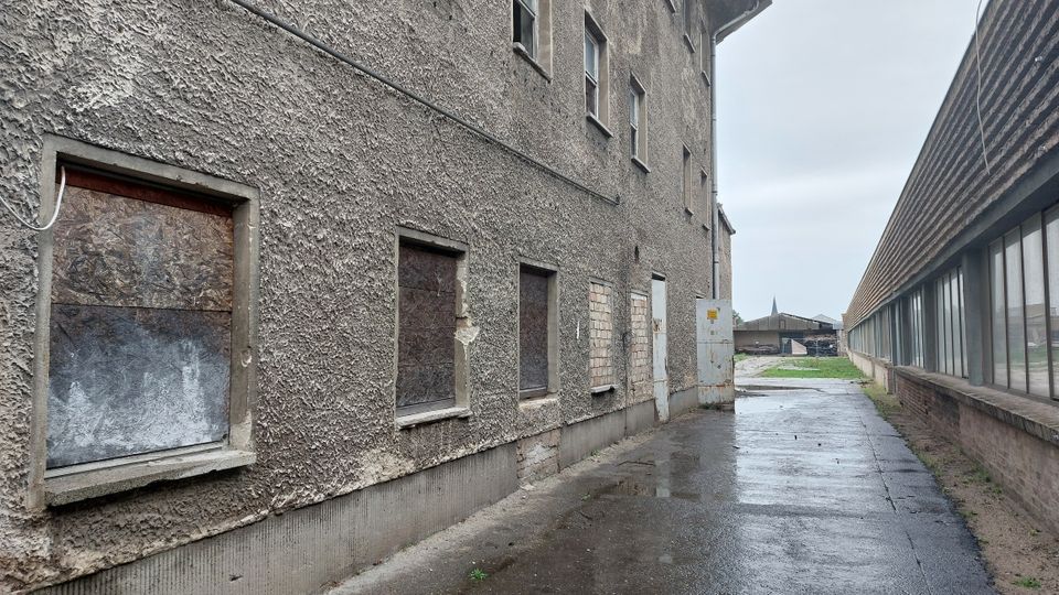 Eine schmale Gasse zwischen einem Gebäude mit grauer, rauer Fassade und teilweise zugemauerten oder mit Holzplatten verschlossenen Fenstern auf der linken Seite und einer langgestreckten Glasfront eines Industriegebäudes auf der rechten Seite. Der Boden ist regennass, und im Hintergrund sind Höfe, kleine Gebäude und eine Kirchturmspitze sichtbar.
