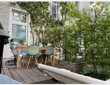 Kleiner Hinterhof mit einer Holzterrasse, darauf ein Esstisch mit bunten Stühlen. Im Hintergrund ist eine mit Efeu bewachsene Hausfassade mit Fenstern, umgeben von Sträuchern und Pflanzen.