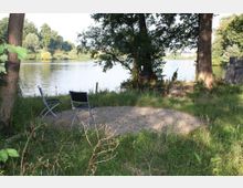 Zwei leere Klappstühle stehen auf einer kleinen Kiesfläche am Ufer eines Sees, umgeben von Gras und hohen Bäumen. Im Hintergrund sind das ruhige Wasser und dicht bewachsene Uferbereiche zu sehen.