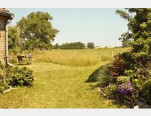 Ein Garten mit Blick auf eine weitläufige Wiese und einzelne Bäume am Horizont. Im Vordergrund befinden sich Blumenbeete, ein kleiner Tisch mit Stühlen und ein Teil einer Hausfassade aus Ziegeln links im Bild.