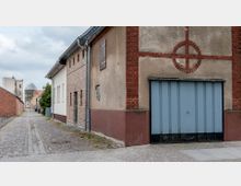 Schmale gepflasterte Gasse mit historischen Gebäuden aus Backstein und Putzfassaden, rechts eine Garage mit hellblauem Tor und Zierelementen aus rotem Backstein. Im Hintergrund sind moderne und ältere Häuser sowie Bäume sichtbar.