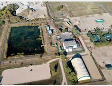 Luftaufnahme eines Reit- oder Freizeitgeländes mit mehreren Sandplätzen, einer großen Halle mit grün-weißem Dach, Stallgebäuden und einem angrenzenden Teich. Umgeben von Wiesen, Wegen und wenigen Bäumen.