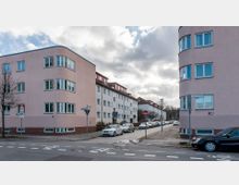 Straßenszene in einem Wohngebiet mit mehrstöckigen Gebäuden in pastellrosa und weißer Farbe, abgerundeten Ecken und Fenstern. Im Vordergrund befinden sich parkende Autos, Straßenschilder und ein Zebrastreifen.