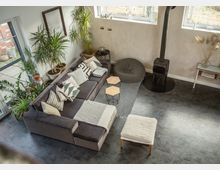 Wohnzimmer in modernem Einrichtungsstil mit einer grauen Eckcouch, mehreren Dekokissen, einem Teppich und einem schwarzen Kaminofen. Im Raum befinden sich zahlreiche Pflanzen, kleine Beistelltische und große Fenster, die viel Tageslicht hereinlassen.