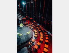 Runde, erhöhte Bühne mit zwei grünen Stühlen und transparenten Podest-Tischen, umgeben von halbkreisförmig angeordneten Stuhlreihen aus Holz und Metall in einem dunklen Raum mit deckenhohen Glasfenstern und roter Beleuchtung.